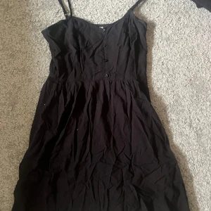 Svart långklänning med knappar - Svart långklänning med smala axelband och knappar framtill. Klänningen har en enkel och stilren design med lätt utställd kjol. Perfekt för varma dagar eller att styla med en jacka. Materialet känns mjukt och lätt.