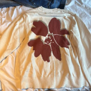 Beige stickad tröja Jack & Jones M - Beige stickad tröja från Jack & Jones med stor blommig design i vinrött och grått på ryggen. Rund halsringning och ribbade muddar. Perfekt för dig som gillar unika detaljer och vill sticka ut lite extra.