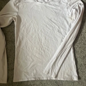 Vit långärmad basictröja i bomull - Enkel och stilren vit långärmad tröja i mjuk bomull. Perfekt som basplagg och funkar till allt. Tröjan har rund halsringning och en normal passform som gör den lätt att matcha med andra plagg. Är missfärgad under armhålan se bild