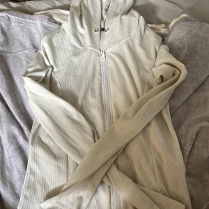 Vit randig tröja med huva - Ljusvit vindjacka med tunna vertikala ränder och hel dragkedja framtill. Jackan har huva med tryckknappar och är tillverkad i ett lätt, ribbat material. Perfekt för vår eller sommarkvällar när du vill ha något enkelt och stilrent.