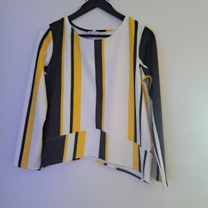 Randig långärmad topp i gult och svart - Snygg långärmad topp från Blanco med breda vertikala ränder i vitt, gult och svart. Modellen har rund halsringning, asymmetrisk nederkant och dragkedja i sidan. Perfekt för dig som gillar färg och grafiska mönster.