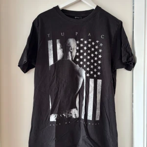 Svart Tupac t-shirt med tryck - Svart t-shirt med stort Tupac-tryck på framsidan och amerikansk flagga i grått och vitt. Klassisk rund hals och korta ärmar. Materialet är mjuk bomull och passformen är normal. Perfekt för dig som gillar streetstyle och ikoniska prints.