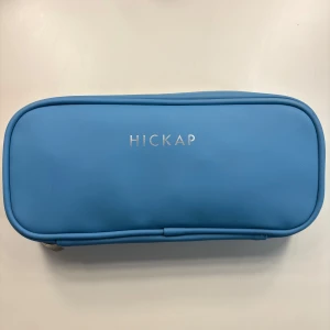 Blå necessär från Hickap - Säljer en stilren blå necessär från Hickap med rosa insida och flera meshfickor. Väskan är avlång och har dragkedja runtom, perfekt för att organisera smink eller småprylar. Utsidan är slät och enkel med diskret logga i silver. Meshfickorna är lite smutsiga av smink men bör gå att få bort.