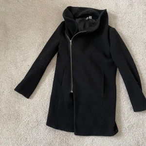 Svart kappa med dragkedja - Snygg svart kappa med asymmetrisk dragkedja och hög krage. Perfekt för kyliga dagar med sin stilrena design och långa ärmar. En tidlös klassiker som passar till det mesta.