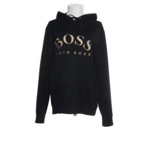 Svart hoodie från Hugo Boss - Snygg svart hoodie från Hugo Boss med guldtryck på bröstet. Perfekt för en stilren och bekväm look. Den har långa ärmar och en klassisk huva med dragsko.