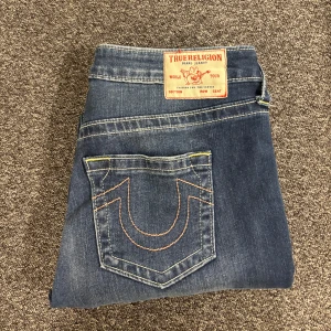 Blå jeans från True Religion - Snygga blå jeans från True Religion med klassisk femficksdesign och kontrastsömmar. De har en bootcut-stil och är perfekta för en avslappnad look. Märkeslappen på baksidan ger en extra touch av stil.