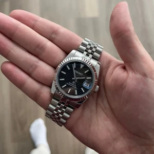 Seiko Datejust Mod Svart - Seiko Mod Datejust är inspirerad av den ikoniska Datejust-designen och drivs av Seiko NH35 automatisk rörelse. En 36/39 mm klocka som kombinerar elegans och tillgänglighet.  Funktioner: • Diameter: 36 eller 39 mm • Rörelse: Seiko NH35 automatisk • Tjocklek: 12 mm • Glas: Safir, rep-resistent • Luminova: Ja • Material: 316L rostfritt stål • Armband: 316L stål med säkerhetslås • Bakstycke: Kristall (visar drivverk) • Vattentålig: 3 ATM