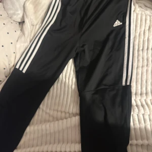 Svarta träningsbyxor från Adidas - Snygga svarta träningsbyxor från Adidas med de klassiska tre vita ränderna längs sidorna. Byxorna har en elastisk midja för extra komfort och är perfekta för sport och träning. Passar mig perfekt som bär storlek S 
