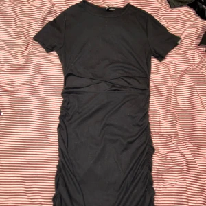 Svart klänning från Divided - Snygg svart klänning från Divided med korta ärmar och en tight passform. Klänningen har en omlott-detalj i midjan som ger en fin siluett. Perfekt för en stilren look.