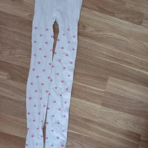Vita strumpbyxor med rosa hjärtan - Säljer ett par vita strumpbyxor dekorerade med små rosa hjärtan över hela ytan. Perfekta för att ge en söt touch till din outfit. Strumpbyxorna är stretchiga och bekväma att bära.