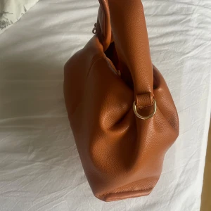 Brun handväska  - Snygg brun handväska i skinn med en stilren design. Väskan har en praktisk storlek och är perfekt för att bära med sig det viktigaste. Den har en enkel och elegant look med rundade handtag.