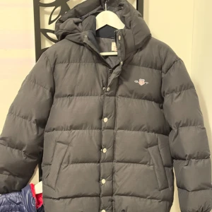 RELAXED PUFFER JACKET - KILLE 146-152  - Svart vadderad jacka från Gant. Vaddering är 100% polyester. Jackan har en avtagbar huva och två fickor med knapp i sidan. På bröstet är märkets välkända logga placerad. Detta är en klassisk jacka som funkar lika bra år efter år.  Jacka  Huva (avtagbar)  Vadderad med polyester  Dragkedja  Knappar  Resår  Framfickor med knapp  Tryck  Färg: 5 Black 