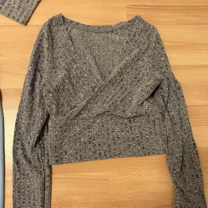 Grå ribbad omlottopp - Säljer en snygg grå ribbad omlottopp med långärmade ärmar. Perfekt för en stilren look. Toppen har en omlottdesign som ger en fin passform. Passar bra till både jeans och kjol.
