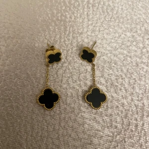 Guldörhängen i rostfrittstål med svart klöverdesign - Eleganta guldörhängen med en svart klöverformad design. Örhängena har en hängande del som ger en stilfull look. Perfekta för att ge en touch av lyx till din outfit. Rostar ej, helt nya