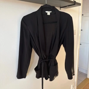 Svart kavaj med knytband - Elegant svart kavaj från H&M med knytband i midjan. Den har en stilren design med långa ärmar och en öppen front. Perfekt för en chic look. Tunnt tyg. 