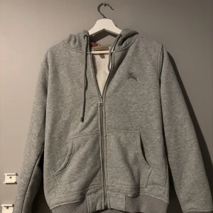 Grå hoodie från Burberry - Säljer en stilren grå hoodie från Burberry med dragkedja och klassiskt rutigt mönster i luvan. Tröjan har långa ärmar och en broderad logga på bröstet. Perfekt för en avslappnad och trendig look.