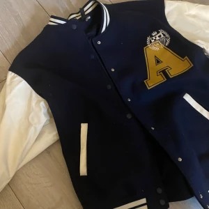 Blå och vit bomberjacka med bokstavsdetalj - Snygg blå och vit bomberjacka med stora bokstaven 'A' i gult på bröstet. Jackan har vita ärmar och ribbade muddar med ränder. Perfekt för en cool och avslappnad stil. Inga defekter, strl S och har passformen oversized☺️