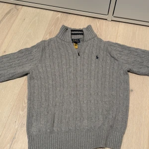 Grå stickad tröja från Polo Ralph Lauren - Säljer en stilren grå stickad tröja från Polo Ralph Lauren med kabelstickat mönster och en liten broderad logga på bröstet. Tröjan har en halv dragkedja vid halsen och ribbade muddar vid ärmslut och nederkant. Perfekt för kyligare dagar! Har andvänr den nån gång bara.