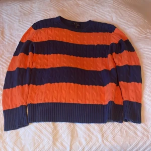 Randig stickad tröja från Ralph Lauren - Säljer en snygg stickad tröja från Ralph Lauren med breda ränder i marinblått och orange. Tröjan har en klassisk design med kabelstickat mönster och långa ärmar. Perfekt för en stilren look!