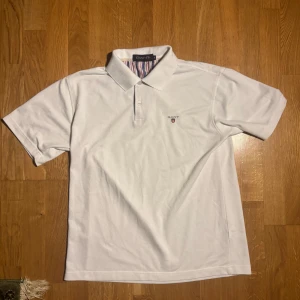 Vit pikétröja från GANT - Säljer en klassisk vit pikétröja från GANT med korta ärmar och broderad logga på bröstet. Tröjan har en stilren krage och knappar framtill. Perfekt för en avslappnad men ändå stilren look. 🕶️