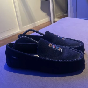 Mörkblå loafers från Polo Ralph Lauren - Snygga mörkblå loafers från Polo Ralph Lauren med broderad björn på ovansidan. Skorna har en klassisk design med mjukt foder för extra komfort. Perfekta för en stilren look.