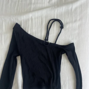 Svart ribbad oneshoulder topp - Snygg svart ribbad topp med oneshoulder-design och lång ärm. Metallring detalj med snören som går över ena axeln. Ena snöretsfäste åkte av så därav en knut där bak som ej märks av. Går att fixa genom att sy fast igen! 🦋🦋