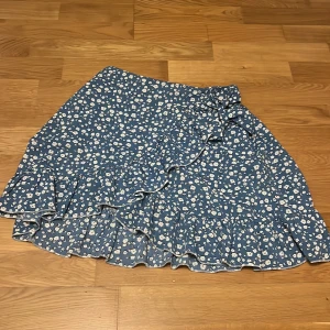 Blå blommig kjol från Shein - Charmig blå kjol från Shein med ett sött blommönster i vitt. Kjolen har volangdetaljer och en omlott-design som ger en lekfull look. Den har även en rosett. 