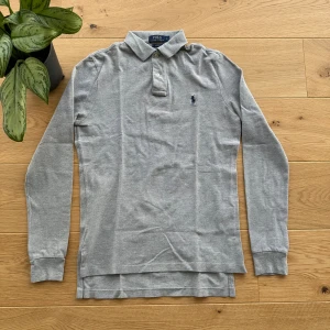 Grå långärmad pikétröja från Ralph Lauren - Säljer en stilren grå långärmad pikétröja från Ralph Lauren. Tröjan har en klassisk krage med knappar och det ikoniska broderade logotypen på bröstet. Perfekt för en avslappnad men ändå elegant look.