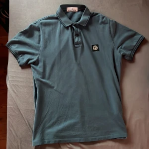 Blå pikétröja från Stone Island - Säljer en stilren blå pikétröja från Stone Island i storlek XL (men passar även stl L). Tröjan har en klassisk krage med knappar och ett broderat Stone Island-märke på bröstet. Perfekt för en avslappnad och snygg look.