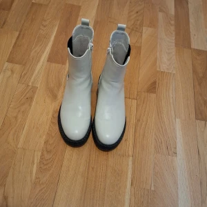 Vita boots med dragkedja - Snygga vita boots i skinn med dragkedja på sidan och svarta detaljer. Perfekta för en stilren look med en robust sula som ger bra grepp. De har en klassisk design som passar till många olika outfits. 