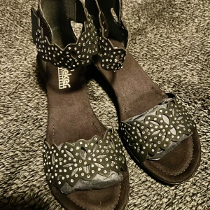 Svarta sandaler med dekorativa detaljer - Snygga svarta sandaler från Rieker med dekorativa utskärningar och glittriga detaljer. Perfekta för att ge en extra touch till din outfit. Sandalerna har en bekväm passform och en stilren design.