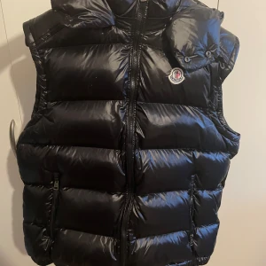 Svart dunväst från Moncler - Snygg svart dunväst från Moncler med dragkedja och hög krage. Perfekt för kyliga dagar när du vill hålla stilen. Västen har Monclers ikoniska logga på bröstet och kommer med en praktisk förvaringspåse.