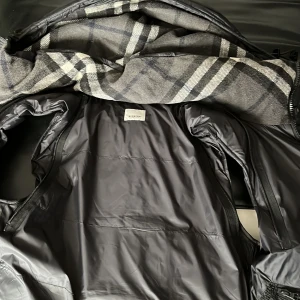 Svart pufferjacka från Burberry - Säljer en snygg svart pufferjacka från Burberry med klassiskt rutigt foder. Jackan har dragkedja och knappar framtill samt praktiska fickor med dragkedja. Perfekt för kyliga dagar! Öppen för byten, pris kan diskuteras