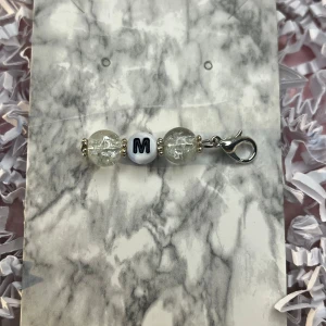 Nyckelring med bokstavspärla - Snygg nyckelring med en vit bokstavspärla 'M' och två genomskinliga glittriga pärlor. Perfekt som personlig accessoar eller present. Har en praktisk karbinhake för enkel fastsättning.