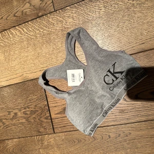 Grå sporttopp från Calvin Klein - Säljer en grå sporttopp från Calvin Klein med deras ikoniska logga tryckt på framsidan. Toppen har en elastisk kant med märkesnamnet runt om och är ärmlös med brottarrygg för extra komfort.