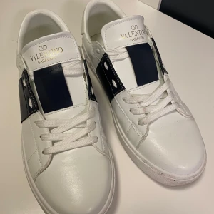 Valentino Garavani sneakers i vitt och blått - Snygga Valentino Garavani sneakers i vitt med blå detaljer. Skorna har en klassisk design med snörning och de ikoniska nitarna runt sulan. Perfekta för en stilren look. Storlek 42 men passar mig som har 43. Angända 2 ggr