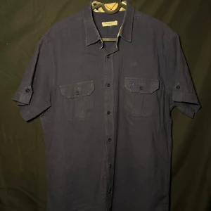 Burberry kortärmad skjorta | Marinblå | Slim/regular | Stl M - Läs nedan för mer info!😊  Snygg mörkblå skjorta från Burberry med korta ärmar och klassisk krage. Skjortan har två bröstfickor med knappar och en diskret broderad logga. Perfekt för en stilren look.  Produktinformation:  ✅ Märke: Burberry ✅ Storlek: M  ✅ Färg: Marinblå ✅ Material: Bomull ✅ Passform: Slim fit – perfekt att styla med jeans eller chinos. ✅ Skick: Mycket bra skick, knappt använd  Frakt och betalning: 📦 Snabb leverans – skickar inom 1-2 dagar efter beställning📦