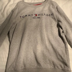 Grå sweatshirt från Tommy Hilfiger  - Säljer en stilren grå sweatshirt från Tommy Hilfiger med deras ikoniska logga och text 'Est. 1985' på framsidan. Tröjan har långa ärmar och en rund halsringning. Perfekt för en avslappnad look. 