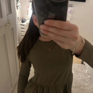 Olivgrön klänning med volangdetalj - Säljer en olivgrön tröja med lång ärm och volangdetalj i midjan. Perfekt för en stilren och bekväm look. Tröjan har en rund halsringning och är gjord i ett mjukt material.