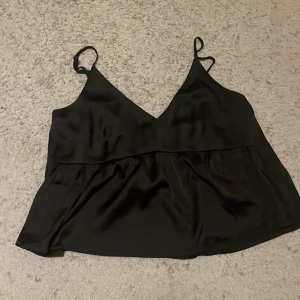 Svart satinlinne - Snyggt svart linne i satin med smala axelband och v-ringning. Perfekt för en stilren look. Den har en defekt som visas på bild 2 men det går lätt att fixa.