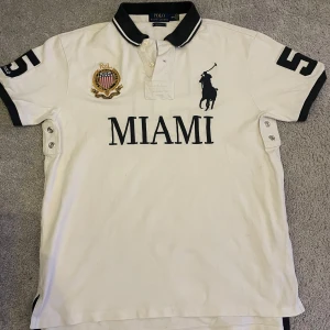 Miami Piké från Ralhp lauren  - Pris är förhandlingsbart!! Säljer en vit pikétröja från Ralph Lauren med svart text 'MIAMI' och en stor siffra '5' på ryggen. Tröjan har korta ärmar och en broderad logotyp på bröstet. Perfekt till sommaren. Tröjan är helt riktig! Nu säljs pikén till ett bra pris!                                      Tröjan har några små fläckar på sig!