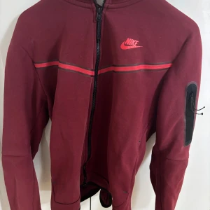 Vinröd jacka från Nike - Säljer en vinröd jacka från Nike med dragkedja och huva. Jackan har en diskret logga på bröstet och en praktisk ficka med dragkedja på ärmen. Perfekt för en sportig look!