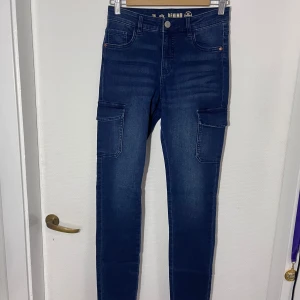 Blå jeans från Elly Denim - Snygga blå cargobyxor från Elly Denim med praktiska fickor på sidorna. Byxorna har en tight passform och är tillverkade i ett bekvämt denimtyg. Perfekta för en stilren och avslappnad look.