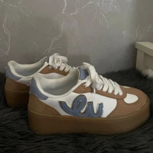 Beige och vita sneakers från Pernilla Wahlgren - Säljer ett par snygga sneakers från Pernilla Wahlgren i beige mocka och vitt syntetmaterial. Skorna har en tjock platt sula, vita snören och blå detalj på sidan. Skorna är alldeles nya & säljs pga felköp 