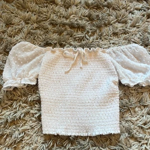 Vit topp  - Supersöt vit topp med smockad kropp och puffiga, korta ärmar i transparent tyg med små broderade prickar. Toppen har knytning framtill och volangdetaljer vid ärmslut och nederkant. Perfekt för en somrig look!