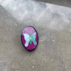 Lila brosch med fjärilsmotiv - Säljer en oval brosch med lila bakgrund och en ljusblå fjäril på framsidan. Broschen har en metallnål på baksidan och är tillverkad på Filippinerna. Perfekt som accessoar för att piffa upp din outfit.