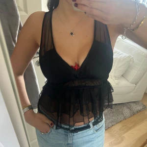 Svart transparent volangblus - Säljer en svart transparent blus med volang och v-ringning. Toppen är ärmlös och har en croppad passform med meshdetaljer. Perfekt att styla med jeans eller kjol.