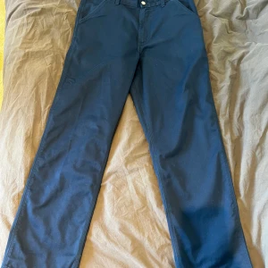 Mörkblå byxor från Carhartt WIP - Säljer ett par mörkblå byxor från Carhartt Work In Progress, modell Simple Pant. Klassisk rak passform med fickor både fram och bak samt Carhartt-logga på bakfickan. Tillverkade i en blandning av polyester och bomull. Mycket bra skick.