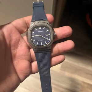 Blå klocka från Dronci - Snygg klocka från Dronci med blått gummiarmband och blå urtavla. Boetten är i silverfärgad metall och har en modern, kantig form. Perfekt accessoar för dig som vill ha en stilren och sportig look.