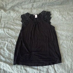 Svart ärmlös topp med spets från H&M - Svart ärmlös topp från H&M med vackra spetsdetaljer på axlarna. Toppen har rund halsringning och är tillverkad i mjukt bomullsmaterial. 